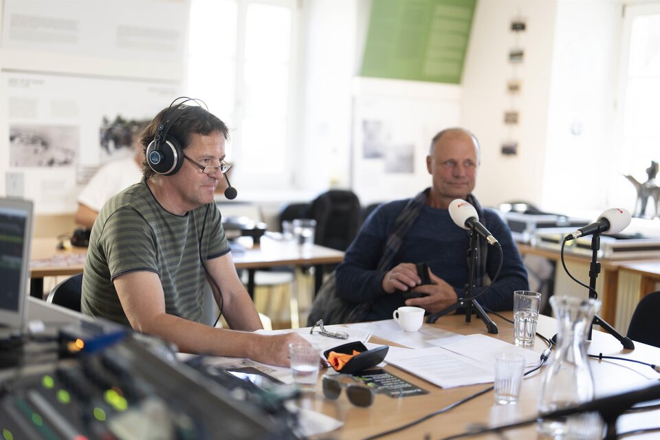 Zwei Männer sitzen an einem Tisch mit Mikrofonen und Kopfhörern. Im Hintergrund sind mehrere Wasserkrüge und Notizen zu sehen. | © Pavelhaus