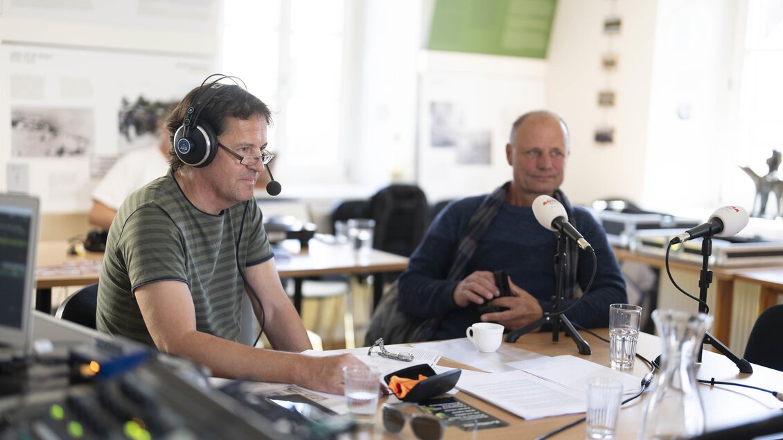 Zwei Männer sitzen an einem Tisch mit Mikrofonen und Kopfhörern. Im Hintergrund sind mehrere Wasserkrüge und Notizen zu sehen. | © Pavelhaus