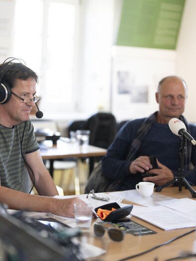 Zwei Männer sitzen an einem Tisch mit Mikrofonen und Kopfhörern. Im Hintergrund sind mehrere Wasserkrüge und Notizen zu sehen. | © Pavelhaus