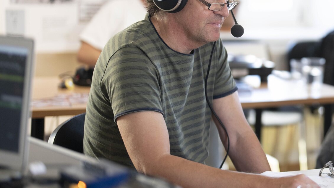 Ein Mann mit Kopfhörern arbeitet an einem Computer. Im Hintergrund sind Tische und weitere Personen sichtbar.