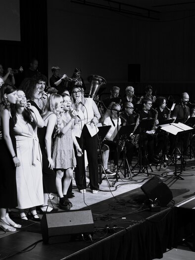 Eine Gruppe von Sängern steht auf der Bühne und gibt ein Konzert. Im Hintergrund ist ein Orchester zu sehen, das die Darbietung begleitet. | © Zehnerhaus