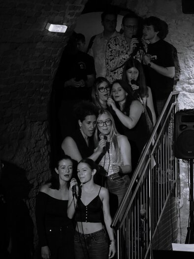 A group of people is passionately singing in a dark room. In the foreground, another person is sitting at the piano. | © Zehnerhaus
