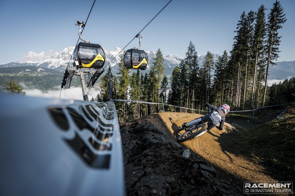 Mountainbiker fährt eine Steilkurve auf einem Downhill-Trail unter einer Gondelbahn mit Bergpanorama. | © Sebastian Gruber