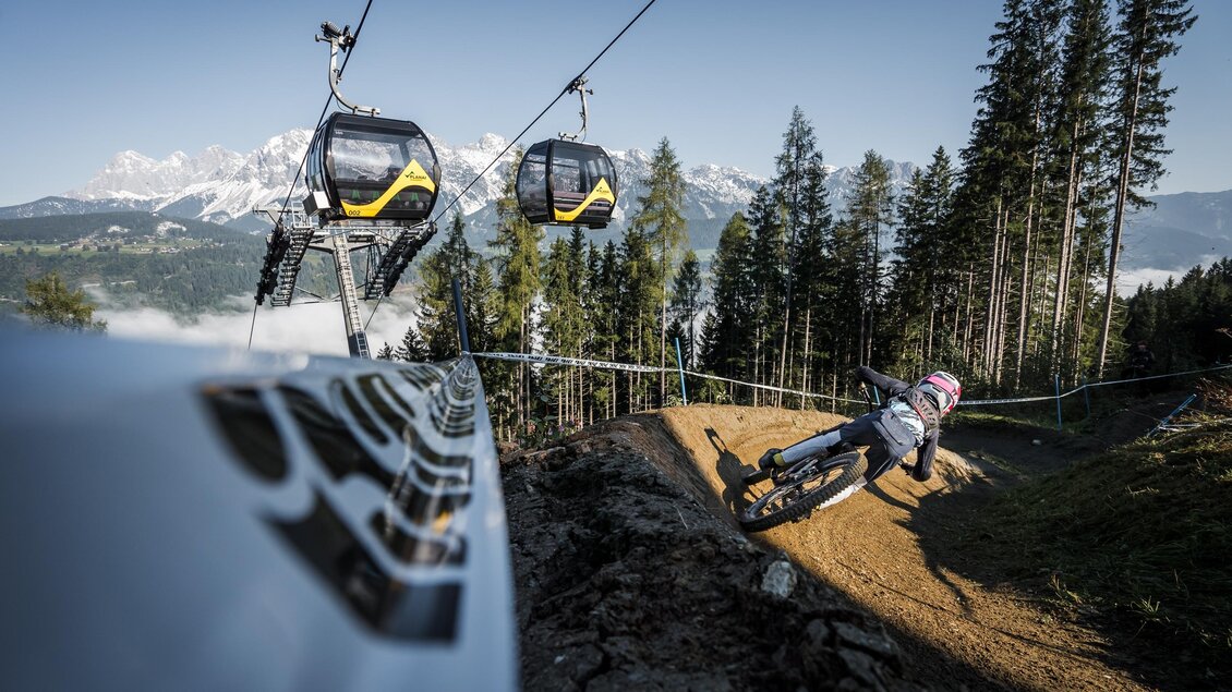 Mountainbiker fährt eine Steilkurve auf einem Downhill-Trail unter einer Gondelbahn mit Bergpanorama. | © Sebastian Gruber