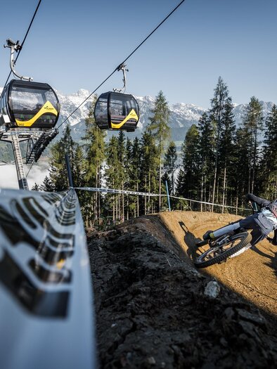Mountain biker rides a steep curve on a downhill trail under a gondola lift with mountain panorama. | © Sebastian Gruber