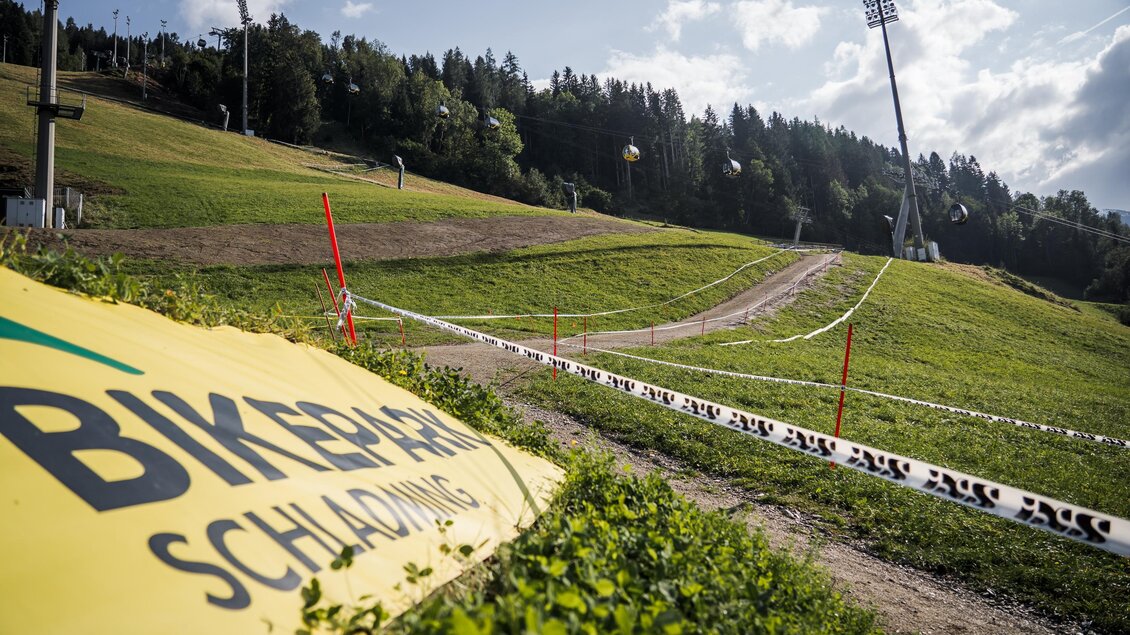 Abgesteckter Bike-Trail auf einer grünen Wiese mit Gondelbahn und Wald im Hintergrund. | © Sebastian Gruber