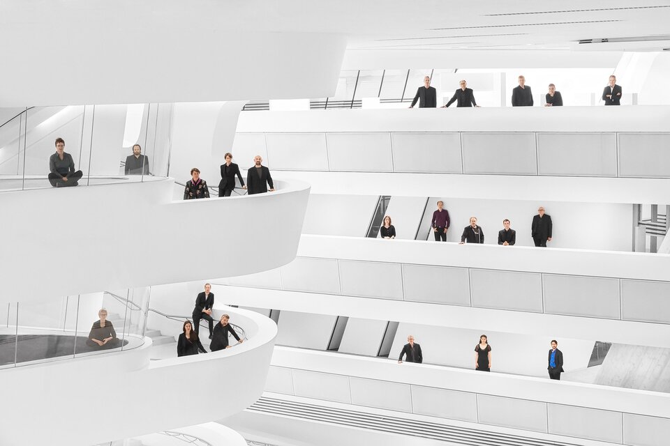 Eine moderne, helle Architektur mit mehreren Ebenen. Menschen stehen auf den Balustraden und in verschiedenen Positionen im Raum. | © _c__Tina_Herz