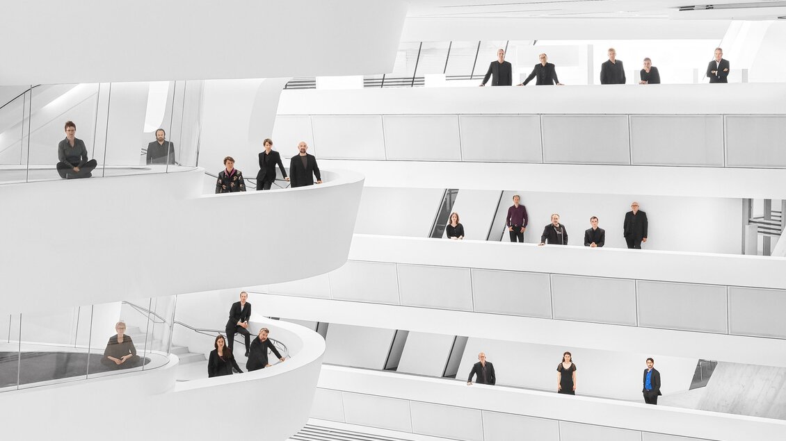 Eine moderne, helle Architektur mit mehreren Ebenen. Menschen stehen auf den Balustraden und in verschiedenen Positionen im Raum. | © _c__Tina_Herz