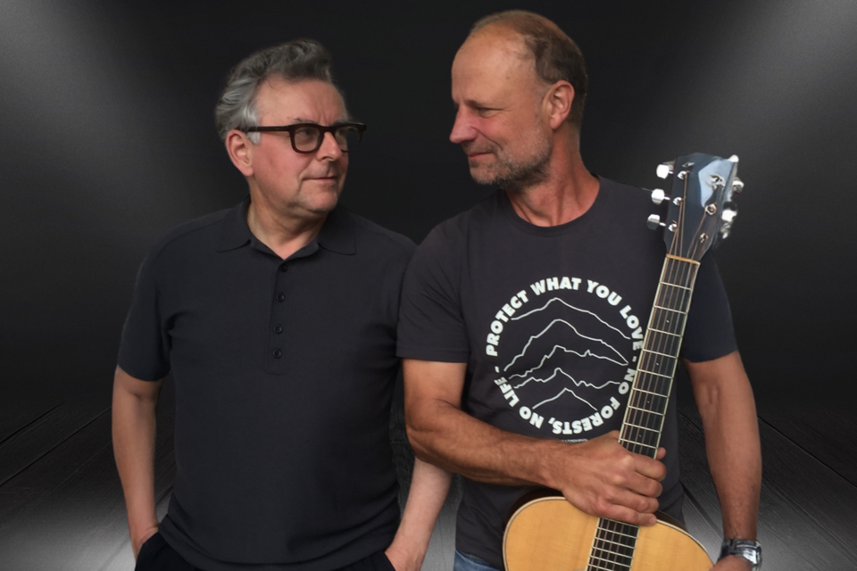 Zwei Männer stehen nebeneinander, einer mit einer Gitarre in der Hand. Der Hintergrund ist dunkel und hebt die Personen hervor. | © Kulturforum