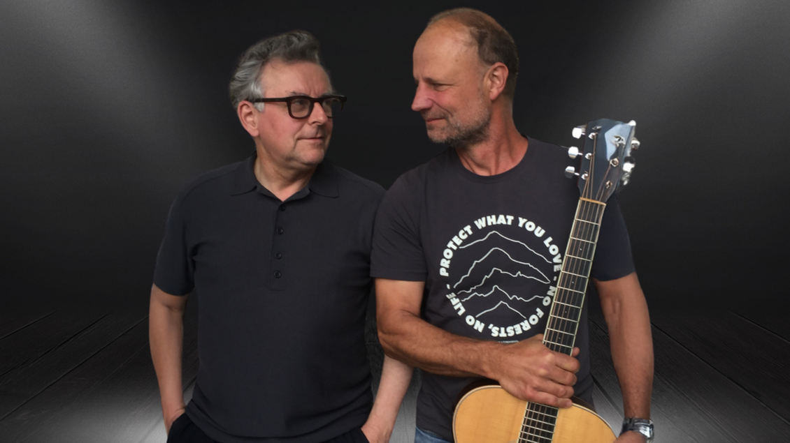 Zwei Männer stehen nebeneinander, einer mit einer Gitarre in der Hand. Der Hintergrund ist dunkel und hebt die Personen hervor. | © Kulturforum