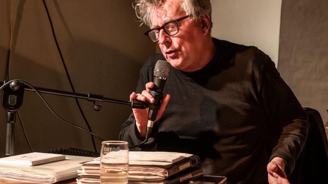 Ein Mann sitzt an einem Tisch und spricht in ein Mikrofon. Vor ihm liegen Bücher und ein Glas Wasser. | © Kulturforum