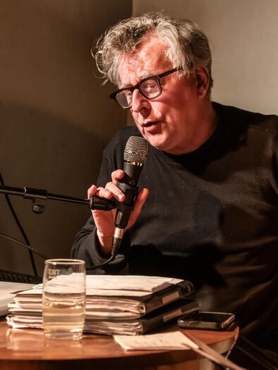 Ein Mann sitzt an einem Tisch und spricht in ein Mikrofon. Vor ihm liegen Bücher und ein Glas Wasser. | © Kulturforum