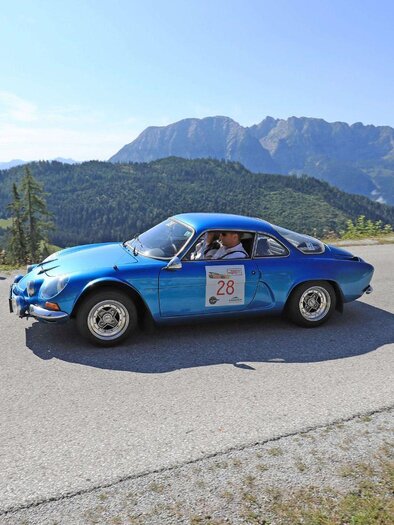 A blue sports car drives along a winding road, surrounded by a picturesque mountain landscape. The sky is clear and the view is stunning. | © Ennstal-Classic_GEPA pictures_Mario Buehner