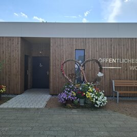 Ein modernes, öffentliches WC mit Holzfassade und bunten Blumenbeeten. Neben dem Eingang steht eine Sitzbank für Besucher. | © Gerlinde Rath