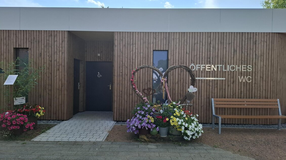 Ein modernes, öffentliches WC mit Holzfassade und bunten Blumenbeeten. Neben dem Eingang steht eine Sitzbank für Besucher. | © Gerlinde Rath