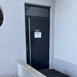 Eine Eingangstür zu einer Toilette mit dem Schild "WC". Die Wand ist hell und die Tür ist dunkel. | © Marktgemeinde Wies
