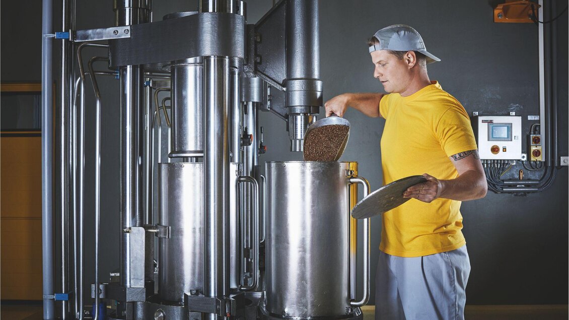 Ein Mann in einem gelben T-Shirt füllt Zutaten in eine große Maschine. Die Szene zeigt eine industrielle Umgebung mit modernen Geräten. | © Ölmühle Fandler