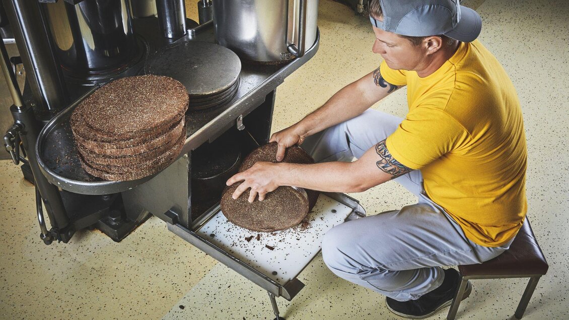Ein Mann in gelber T-Shirt und Kappe arbeitet an einer Maschine, um große runde Platten zu entnehmen. Der Hintergrund zeigt eine industrielle Umgebung. | © Ölmühle Fandler
