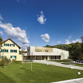 Ein modernes Gebäude neben einem traditionellen Haus in einer ruhigen Landschaft. Der Himmel ist klar und blau mit einigen Wolken, und die Umgebung ist grün und gepflegt. | © Ölmühle Fandler