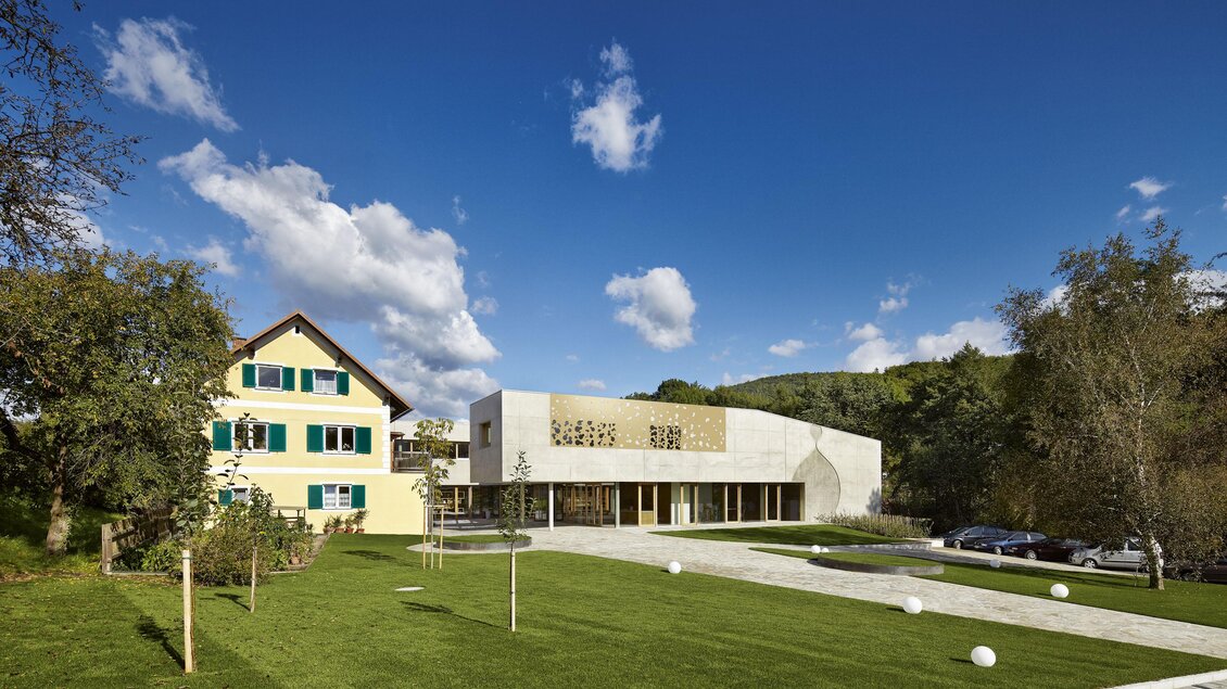 Ein modernes Gebäude neben einem traditionellen Haus in einer ruhigen Landschaft. Der Himmel ist klar und blau mit einigen Wolken, und die Umgebung ist grün und gepflegt. | © Ölmühle Fandler