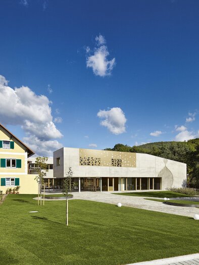 A modern building beside a traditional house in a quiet landscape. The sky is clear and blue with some clouds, and the surroundings are green and well-maintained. | © Ölmühle Fandler