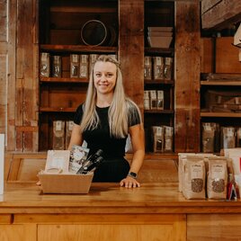 Eine blonde Frau steht hinter einem Holzschalter in einem Laden. Im Hintergrund sind zahlreiche Produkte in Tüten zu sehen. | © Regionalmanagement Südweststeiermark GmbH/webquartier