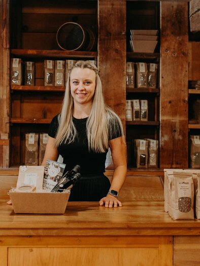 A blonde woman stands behind a wooden counter in a store. In the background, numerous products can be seen in bags. | © Regionalmanagement Südweststeiermark GmbH/webquartier