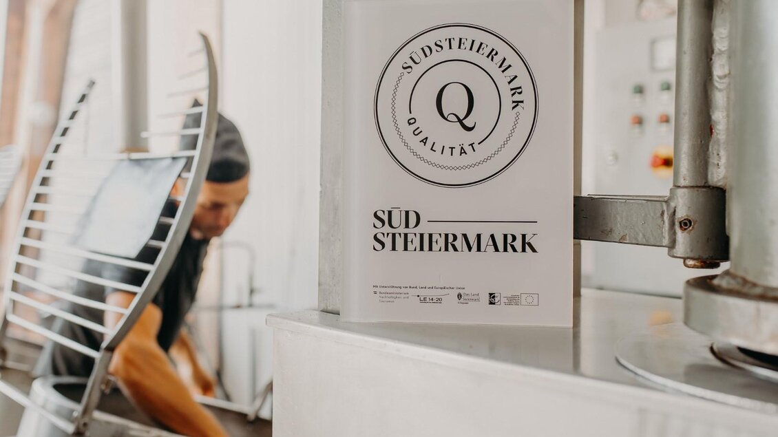 Ein Hinweis auf die Qualität aus der Region Südsteiermark steht im Vordergrund. Im Hintergrund ist ein Koch bei der Zubereitung von Speisen zu sehen. | © Regionalmanagement Südweststeiermark GmbH/webquartier