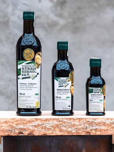 Three bottles of pumpkin seed oil in different sizes are standing on a rustic wooden board. A gray wall is visible in the background. | © RM SW GmbH | webquartier.at