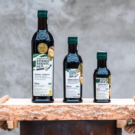 Drei Flaschen Kürbiskernöl in verschiedenen Größen stehen auf einem rustikalen Holzregal. Die Etiketten sind gut sichtbar und zeigen die Produktinformationen. | © RM SW GmbH | webquartier.at