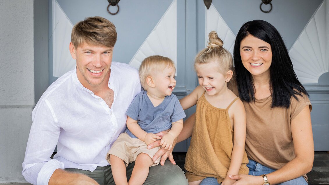 Eine glückliche Familie sitzt zusammen vor einer Haustür. Zwei Kinder lachen, während die Eltern lächeln. | © Pronnegg