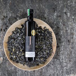 Eine Flasche Kürbiskernöl steht auf einem Teller voller Kürbiskerne. Der Hintergrund ist aus rauem Holz und ergänzt die natürliche Präsentation. | © Pronnegg