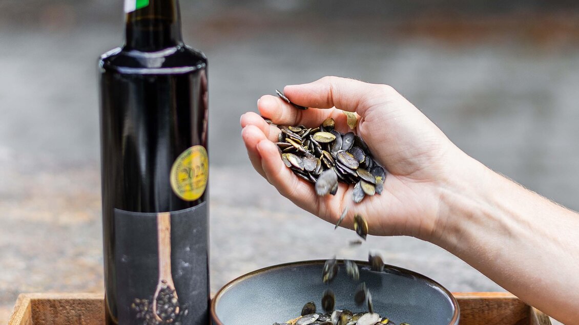 Eine Flasche Öl steht neben einer Schüssel mit Kürbiskernen. Eine Hand hält eine Menge von Kürbiskernen, die gerade aus der Schüssel genommen werden. | © Pronnegg