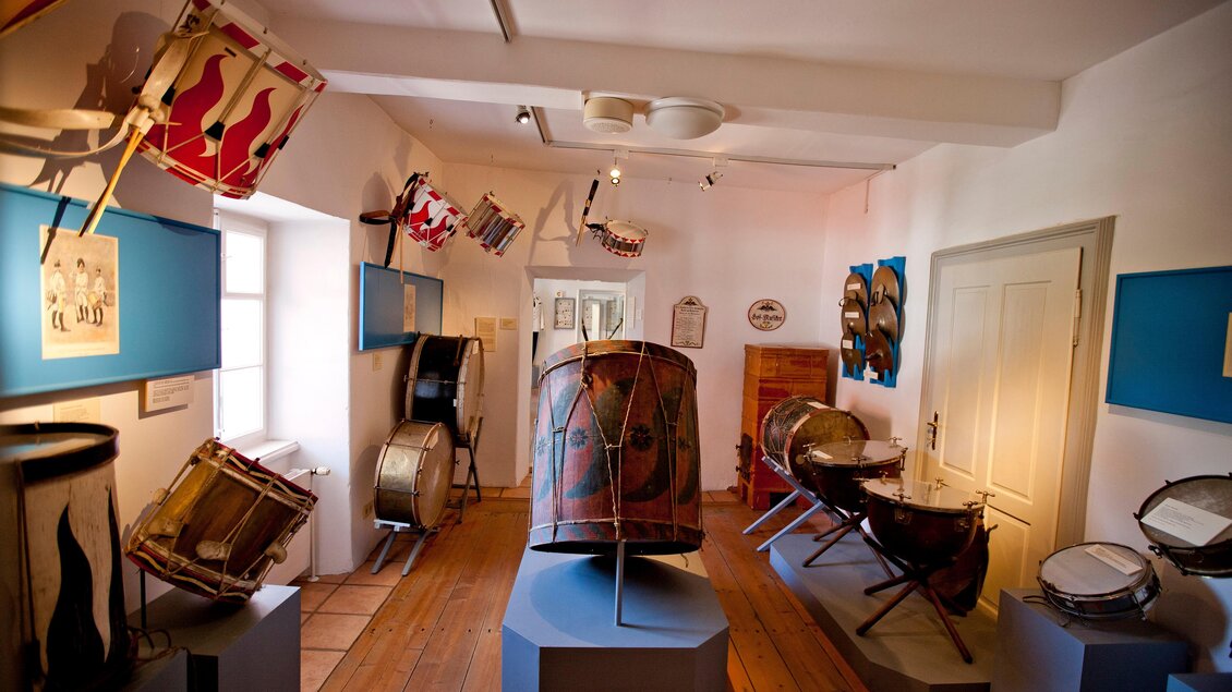 Ein Musikmuseum mit verschiedenen Trommeln und historischen Instrumenten. Die Wände sind mit bunten Fahnen dekoriert. | © Holzwelt Murau