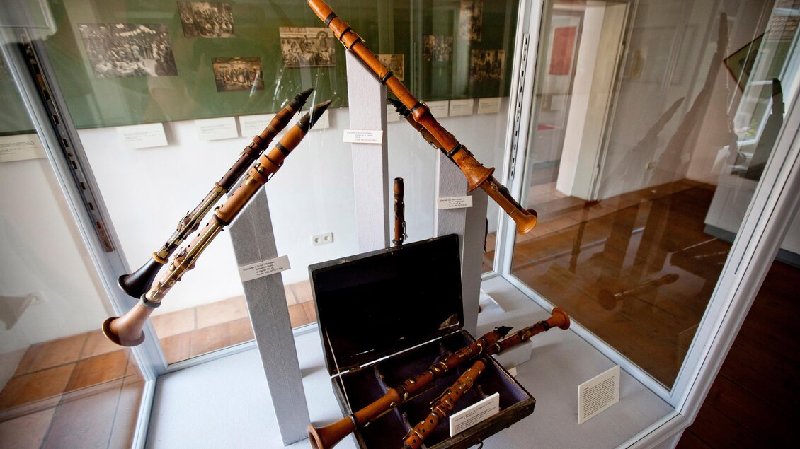 Ein Vitrinenaufsatz mit Holzblasinstrumenten, darunter mehrere Blockflöten und eine elegante Kiste. Der Hintergrund zeigt historische Fotos. | © Holzwelt Murau
