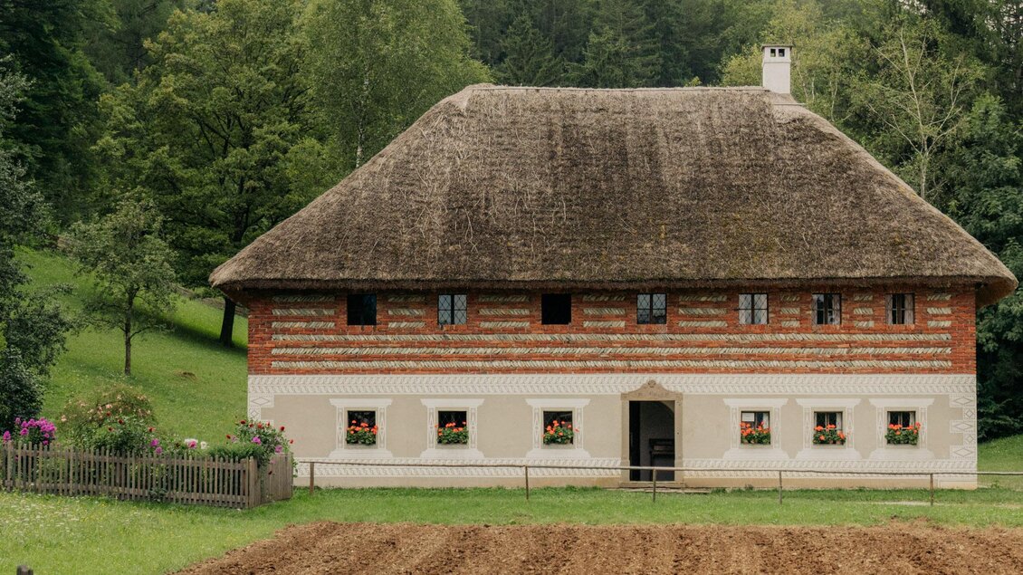 Ein traditionelles Bauernhaus mit einem Reetdach und einem großen Garten. Im Vordergrund ist ein frisch bearbeitetes Feld zu sehen. | © (c) Region Graz - studio draussen