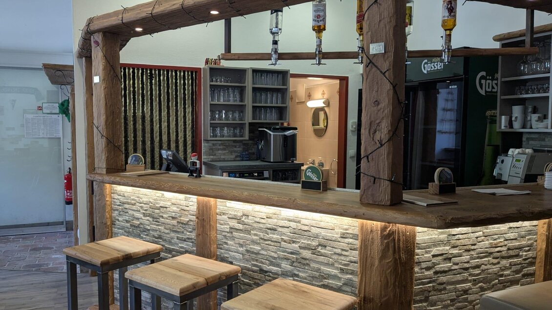 Ein gemütlicher Barbereich mit einem hölzernen Tresen und modernen Hockern. Im Hintergrund sind Regale mit Getränken und einer Kaffeemaschine zu sehen. | © Pub Radmer e.Gen