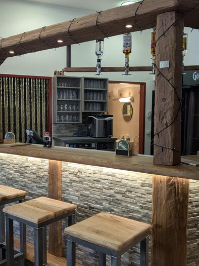 Ein gemütlicher Barbereich mit einem hölzernen Tresen und modernen Hockern. Im Hintergrund sind Regale mit Getränken und einer Kaffeemaschine zu sehen. | © Pub Radmer e.Gen