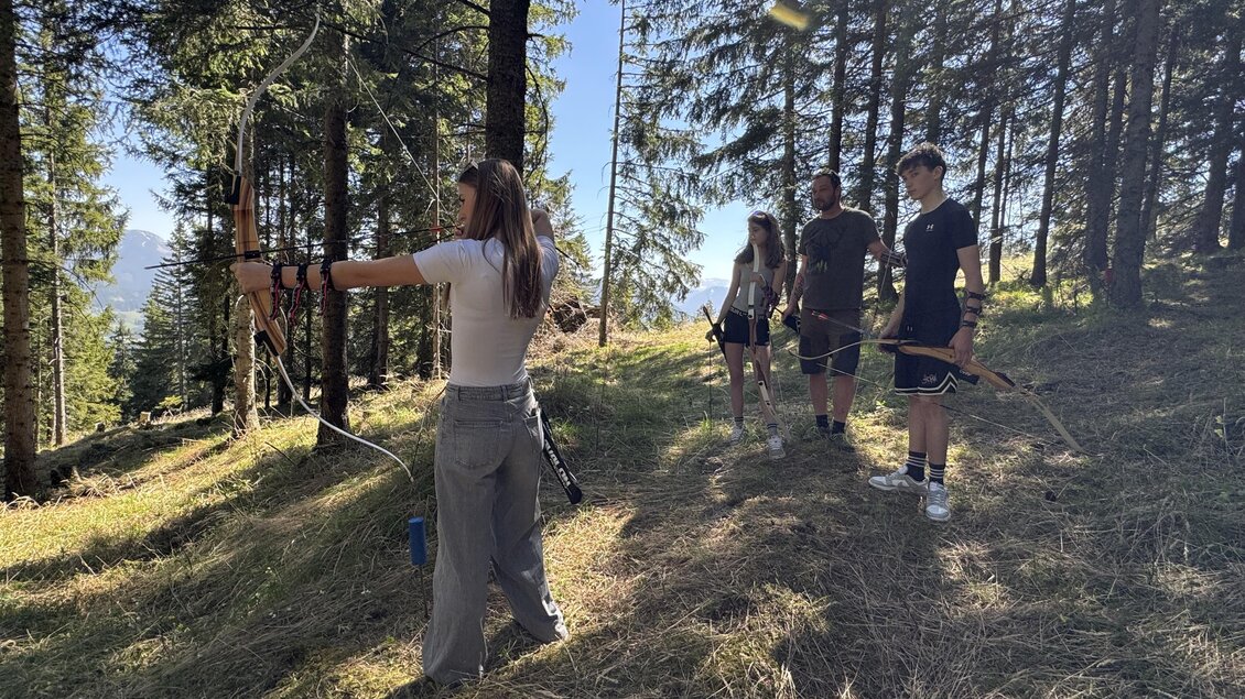 Eine Gruppe junger Leute übt das Bogenschießen im Wald. Die Sonne scheint durch die Bäume und es herrscht eine entspannte Atmosphäre. | © MariazellerBürgeralpe