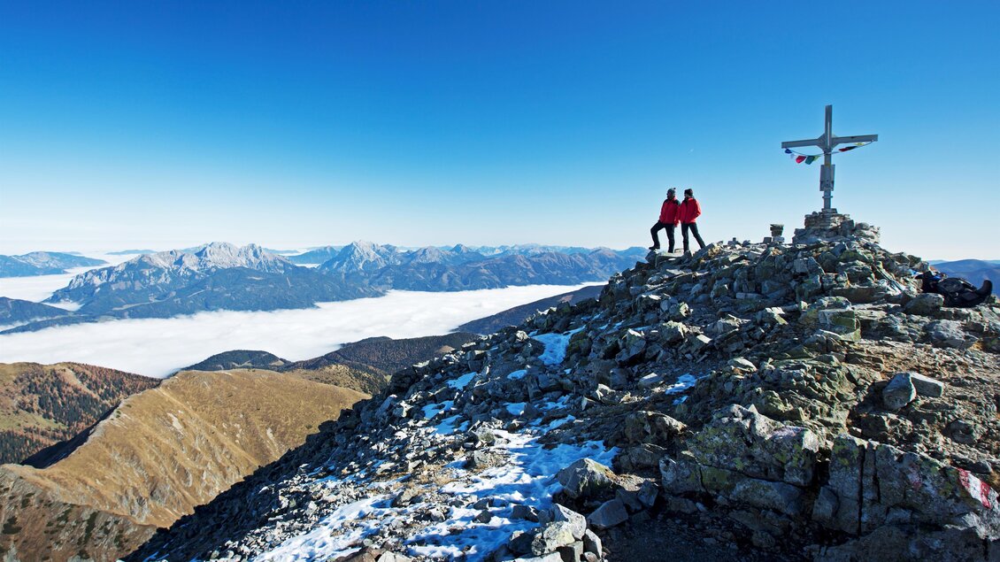Ein Paar steht auf einem Gipfel mit Blick auf eine beeindruckende Berglandschaft. Der Himmel ist klar und blau, und der Boden ist von Schnee und Felsen geprägt. | © Herbert Raffalt