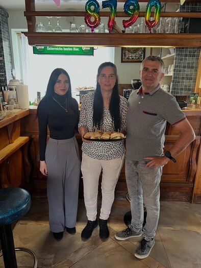 Three people are standing in a restaurant. A woman is holding a tray with food in the middle, while the other two are smiling. | © 8790erPub