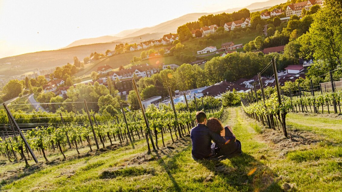 Ein Paar sitzt in einem Weinberg und genießt die Aussicht bei Sonnenuntergang. Die sanften Hügel und das grüne Laub schaffen eine idyllische Atmosphäre. | © Mias Photoart