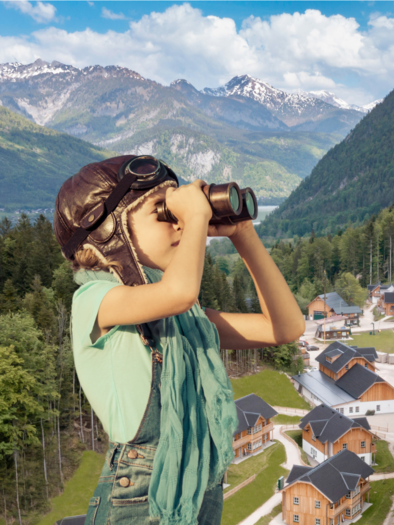 Ein Kind mit einer Fliegerbrille schaut mit einem Fernglas in die Ferne. Im Hintergrund sind Berge und eine kleine, malerische Siedlung zu sehen. | © www.zloam.at