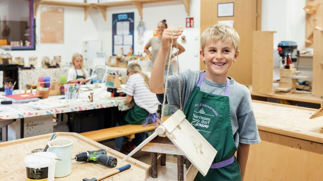 Ein fröhlicher Junge hält ein selbstgebautes Vogelhaus in der Hand. Im Hintergrund arbeiten andere Kinder an verschiedenen Bastelprojekten. | © www.zloam.at