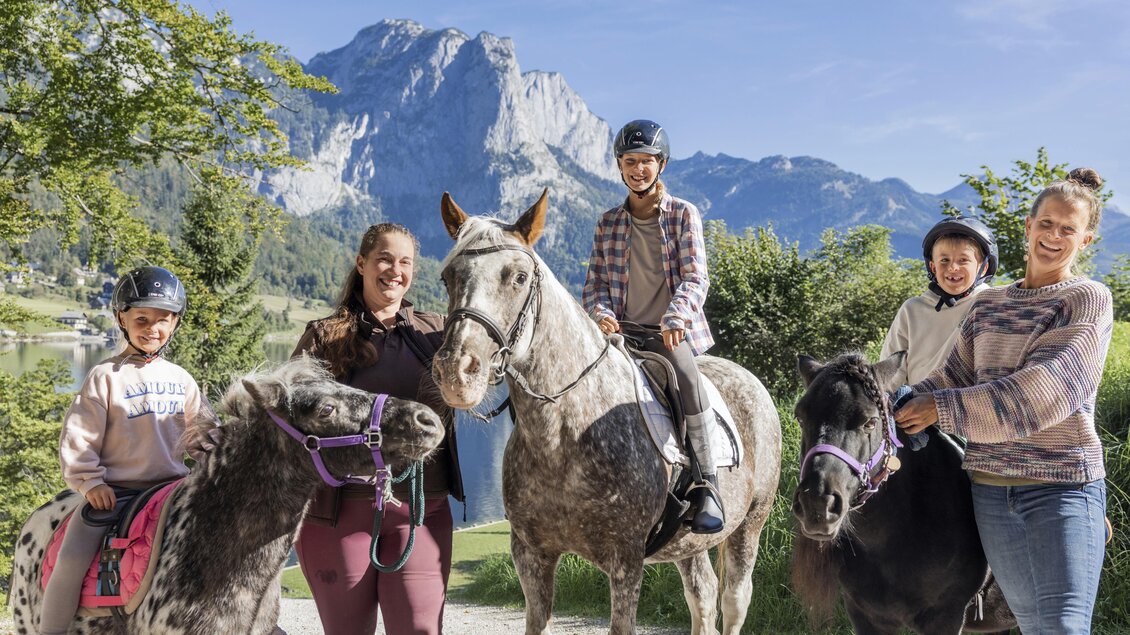 Eine Gruppe von Kindern und Erwachsenen steht mit Pferden in einer schönen Landschaft. Im Hintergrund sind Berge und ein klarer Himmel sichtbar. | © www.zloam.at