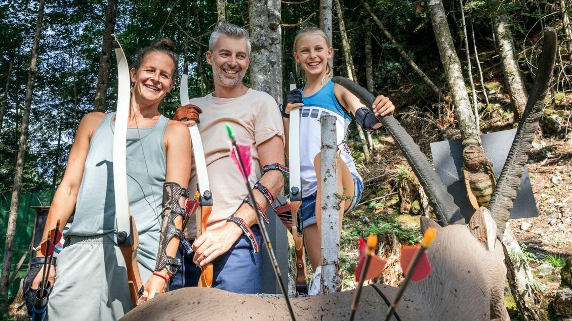 Eine Gruppe von drei Personen steht im Wald und lächelt, während sie mit Bögen und Pfeilen ausgestattet sind. Im Vordergrund ist ein Ziel mit Pfeilen zu sehen. | © www.zloam.at