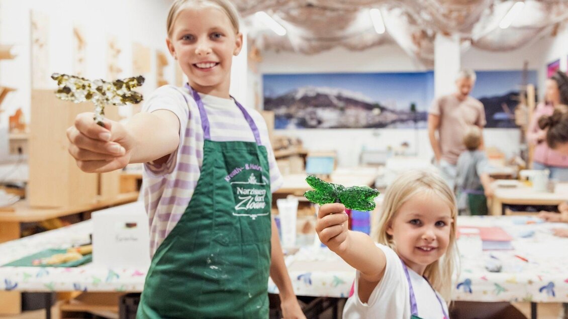 Zwei fröhliche Mädchen halten bemalte Bastelarbeiten in den Händen. Im Hintergrund sind weitere Kinder und ein kreatives Atelier zu sehen. | © www.zloam.at
