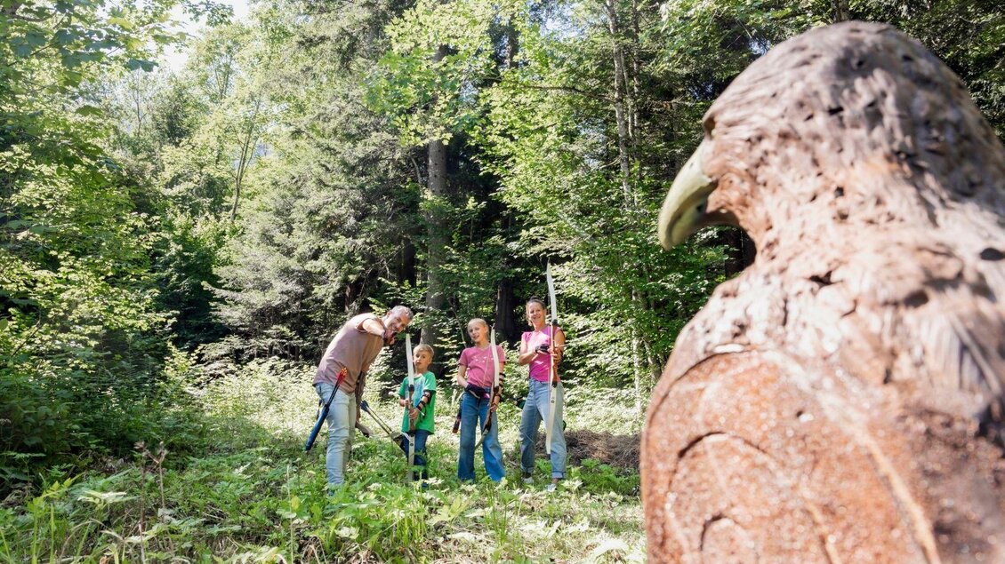 Eine Familie erkundet den Wald und beobachtet einen großen, kunstvollen Vogel aus Holz. Im Hintergrund sind grüne Bäume und sonniges Wetter zu sehen. | © www.zloam.at