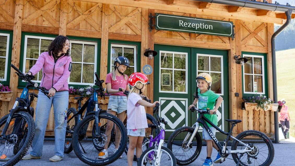 Eine Familie mit Kindern steht vor einem Holzgebäude mit der Aufschrift "Rezeption". Alle tragen Helme und haben Fahrräder dabei, was auf eine Outdoor-Aktivität hindeutet. | © www.zloam.at
