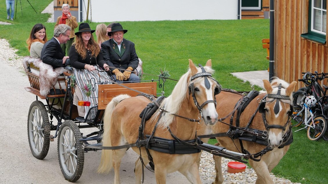 Ein Pferdewagen fährt durch eine malerische Gegend, mit mehreren Personen, die fröhlich darin sitzen. Die Atmosphäre ist ländlich und traditionell, umgeben von Holzhäusern und grünen Wiesen. | © www.zloam.at
